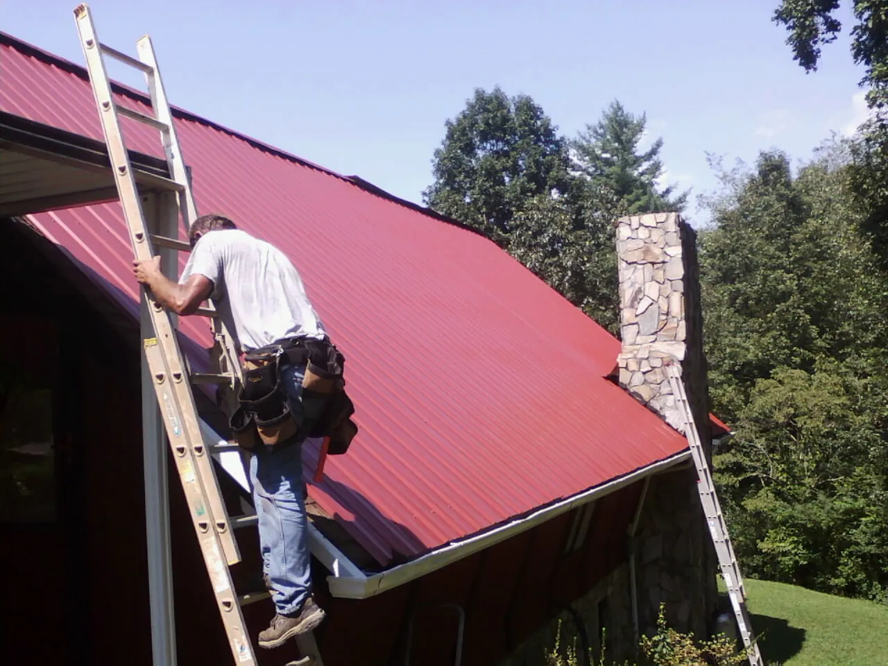Roofer on a ladder beside a red metal roof and stone chimney — Soffit & Fascia Repair by Roofing Brothers in Terrebonne